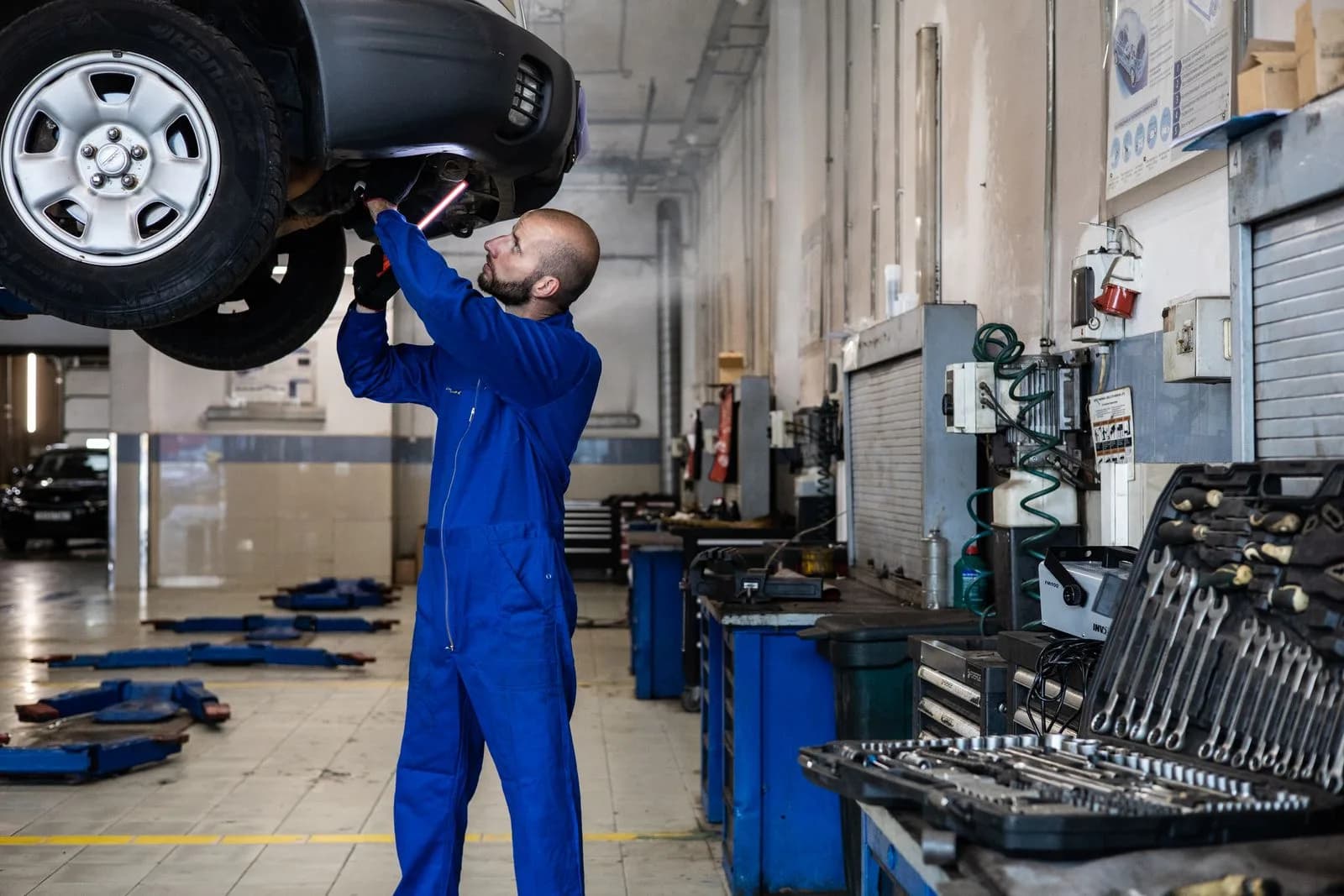A mechanic uses a light to inspect the underside of a raised vehicle.